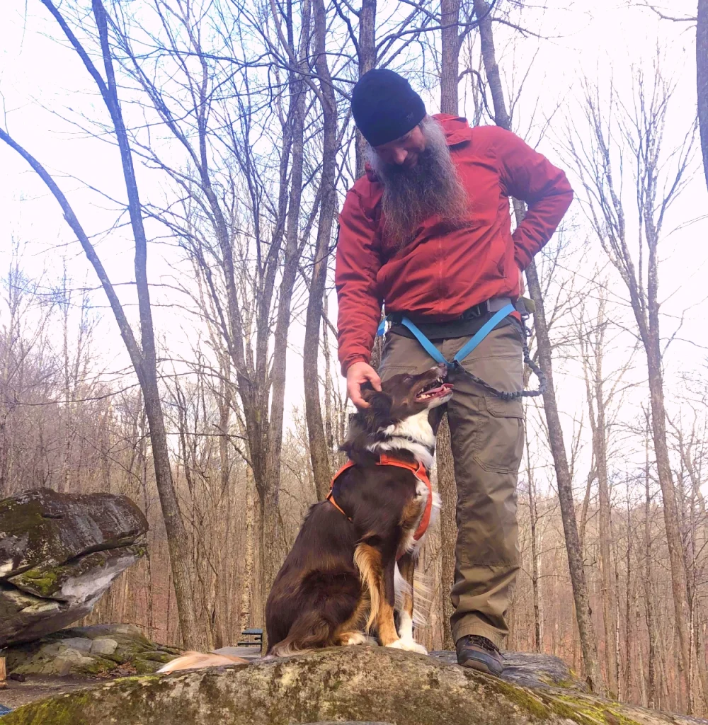Chris Wheeler in hiking gear petting a dog on a rock in a forest.