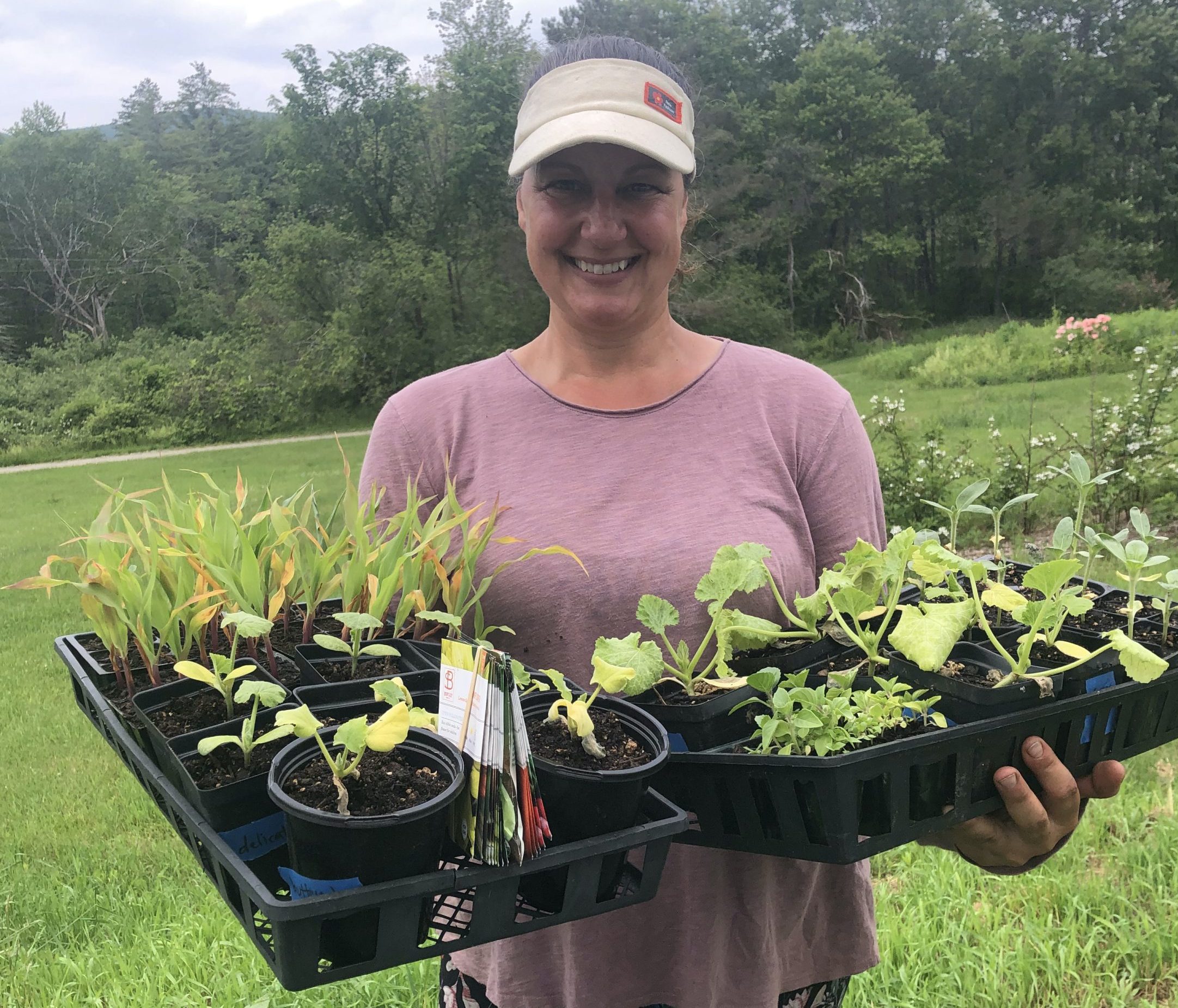 A smiling woman wearing a visor and a pink shirt holding trays of young plants outdoors.