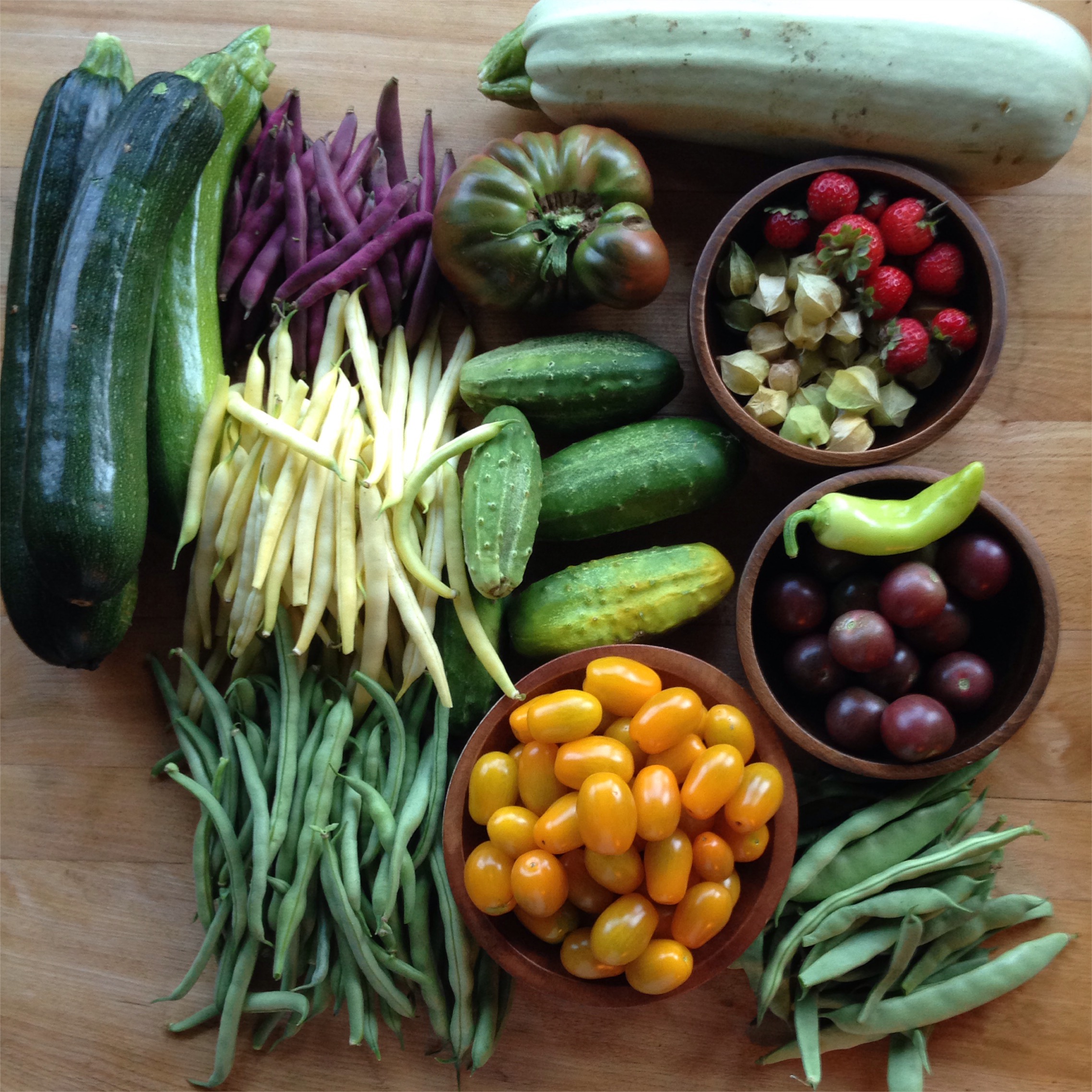 Assorted fresh vegetables and fruits, including zucchinis, beans, cucumbers, cherry tomatoes, strawberries, and ground cherries, arranged on a wooden surface.