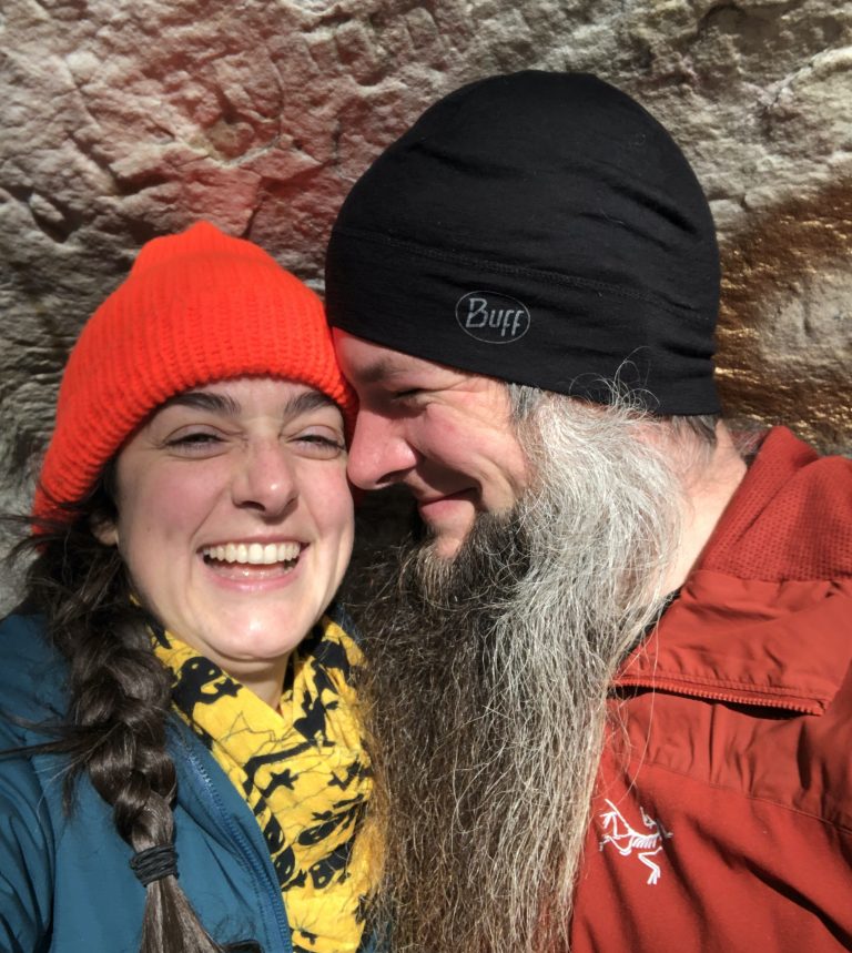Smiling couple wearing winter hats and jackets, standing closely together outdoors.