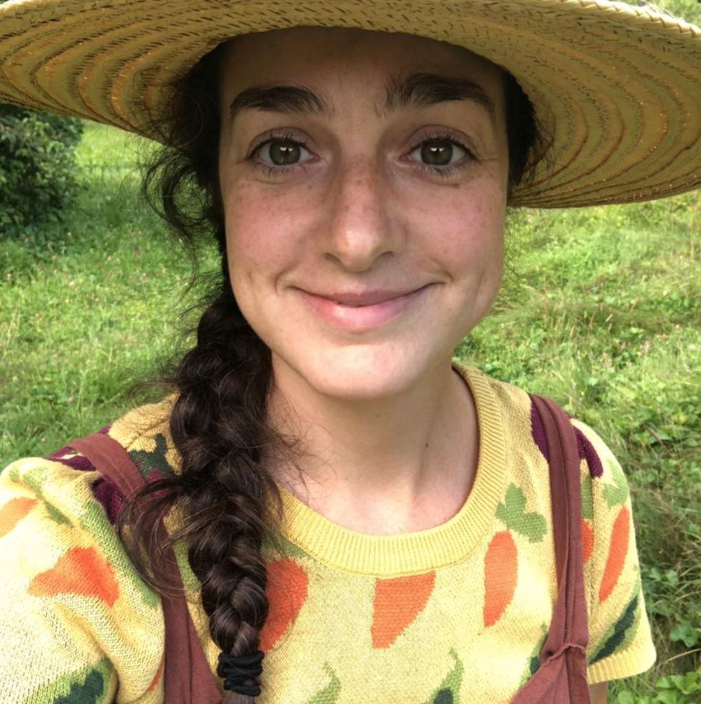 Kristen Tool smiling outdoors wearing a straw hat and a sweater with vegetable patterns.