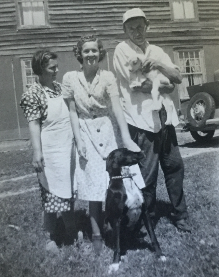 Black-and-white photo of three adults and a dog in front of a house, with one man holding a small animal.