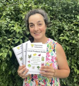 Woman holding three copies of Heart & Soil Collective's "Local Produce Guide" for Berkshire County, Massachusetts, featuring illustrations of vegetables and text promoting local farming in New England.